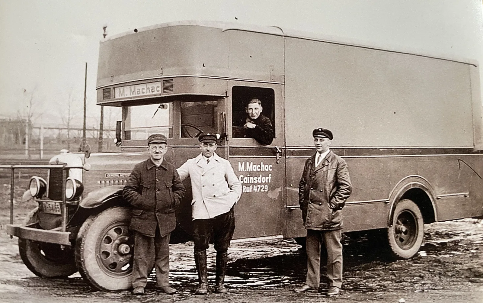 Three men and a driver standing beside a Machac furniture pantechnicon on a muddy yard, the cab lettered “M. Machac, Cainsdorf, Ruf 4729”.